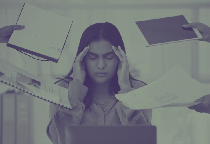 Woman holding her head while working on a laptop with papers flying around, representing workplace stress and overwhelm.